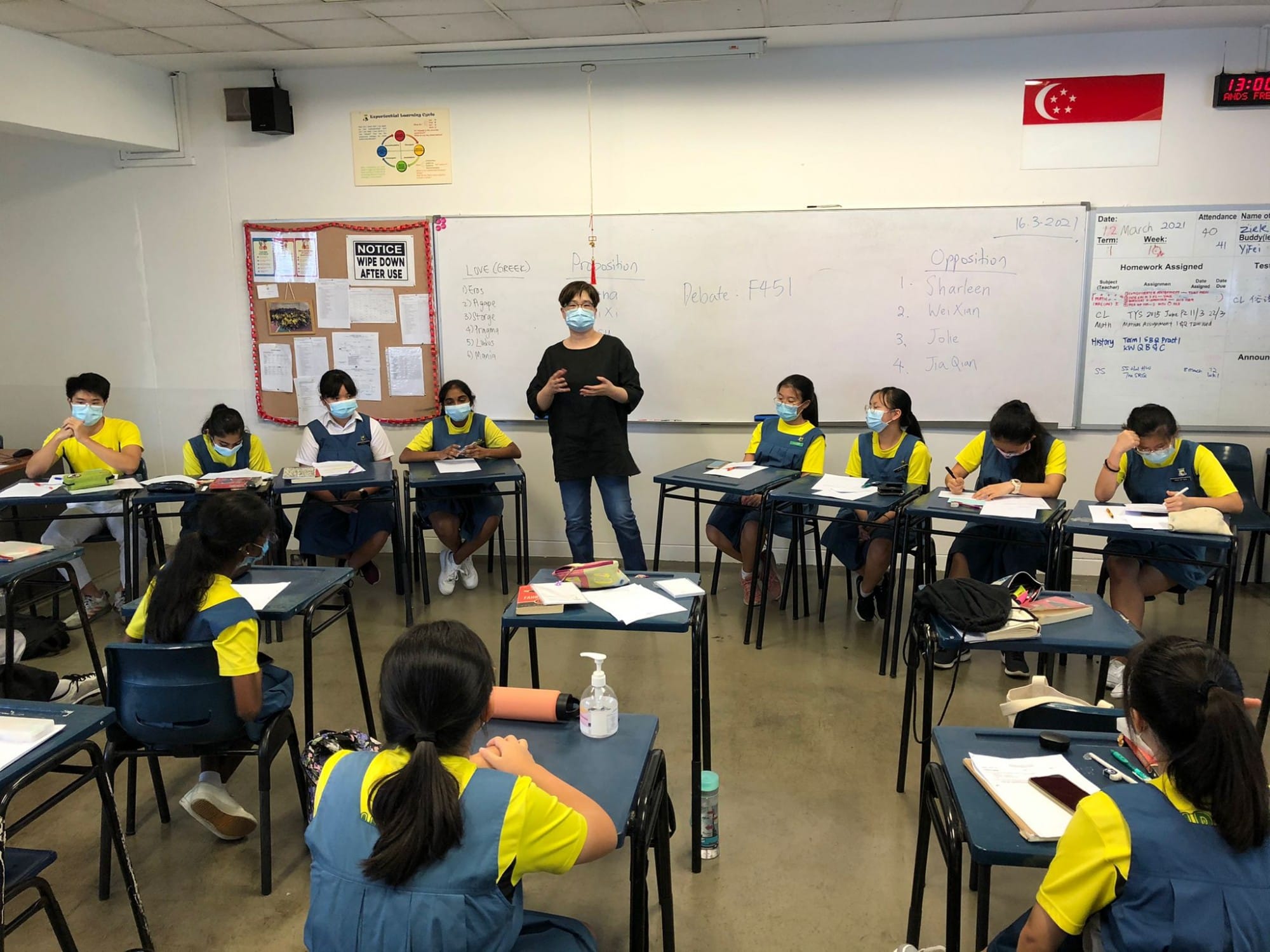 A masked teacher stands before masked students in yellow uniforms seated at desks, with a debate lesson on the whiteboard.